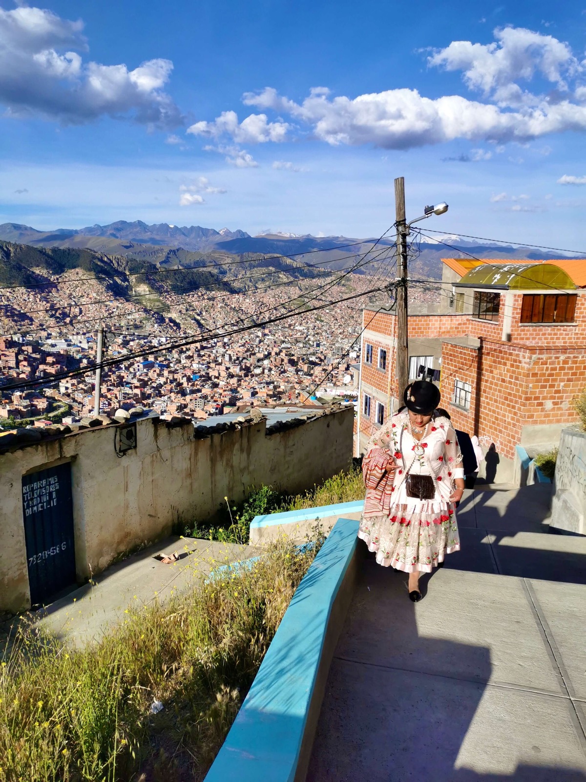Une cholita en tenue traditionnelle marchant dans une ruelle des hauteurs de La Paz, la ville en contrebas
