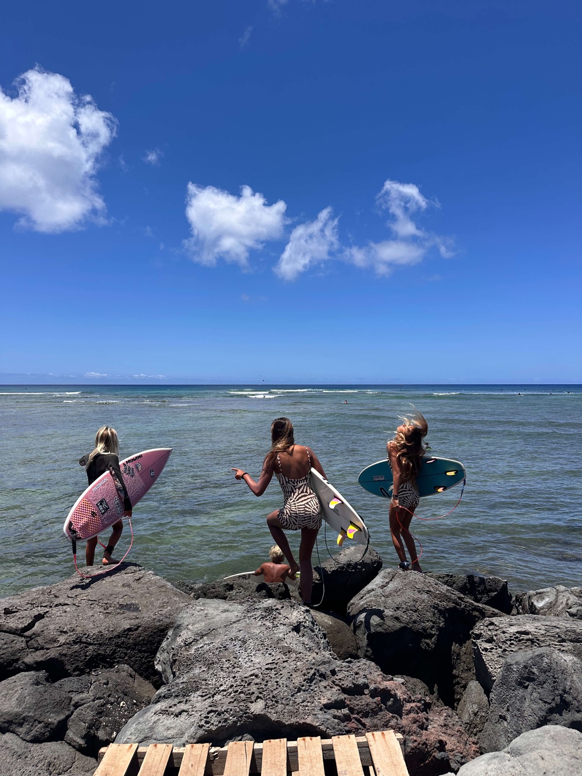 Trois surfeuses descendent vers l'océan, planches sous le bras, sur des rochers volcaniques à Hawaï